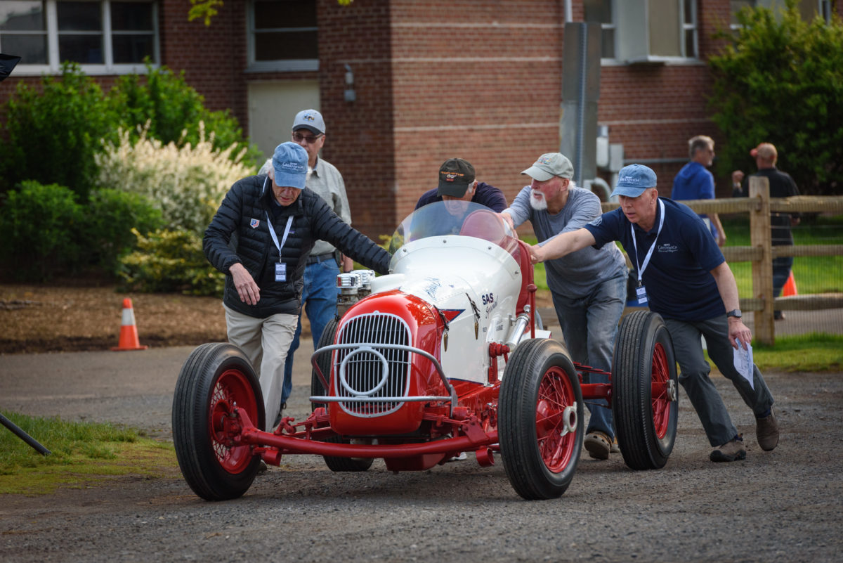 five men in baseball caps pushing red race car down dirt road at Greenwich Concours